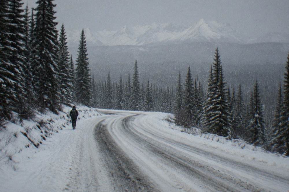 A two-lane snow-packed road curving through spruce forest with mountains in the distance
