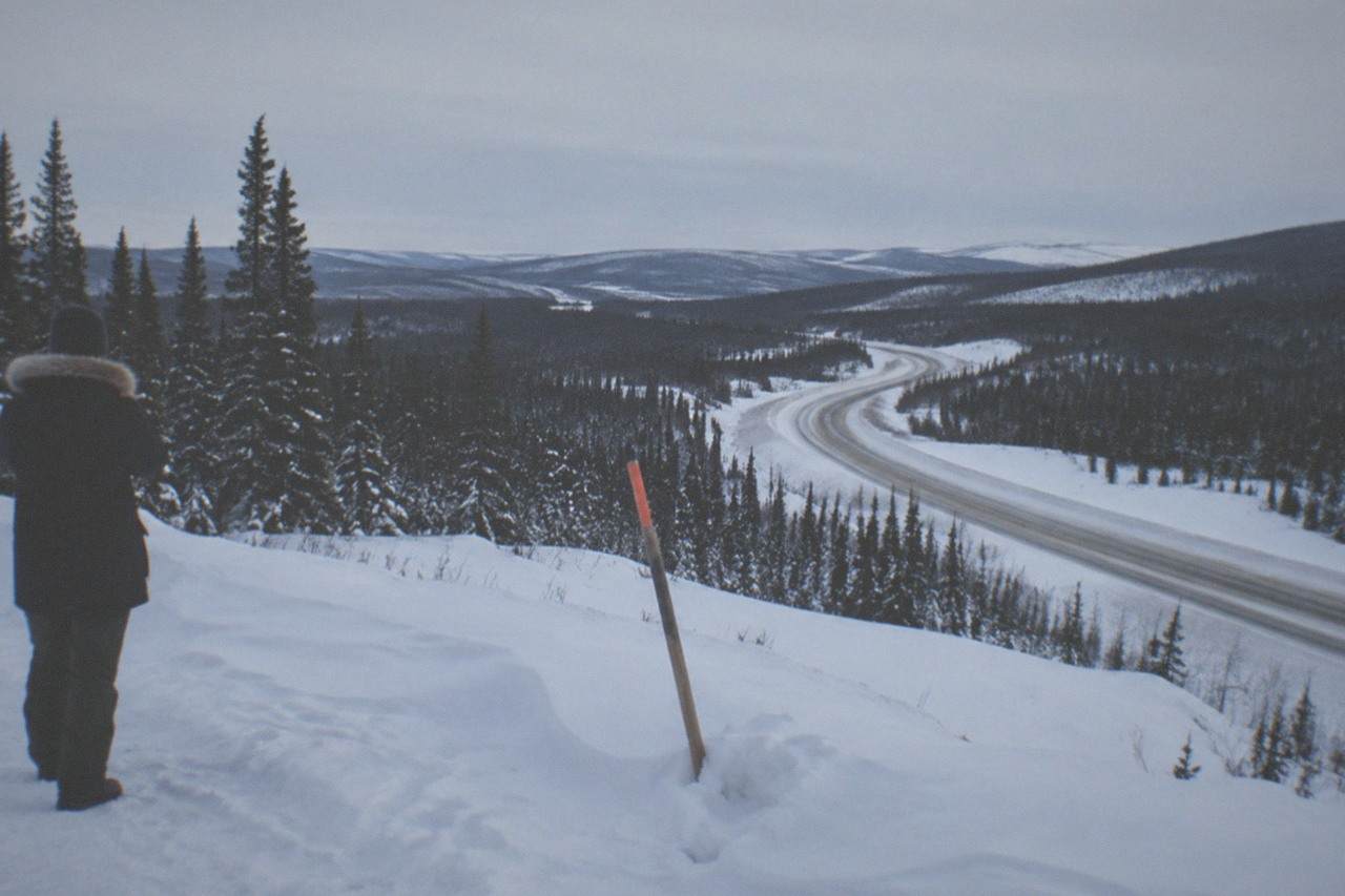 A snow-lined highway corridor through forested Interior Alaska hills under a pale winter sky