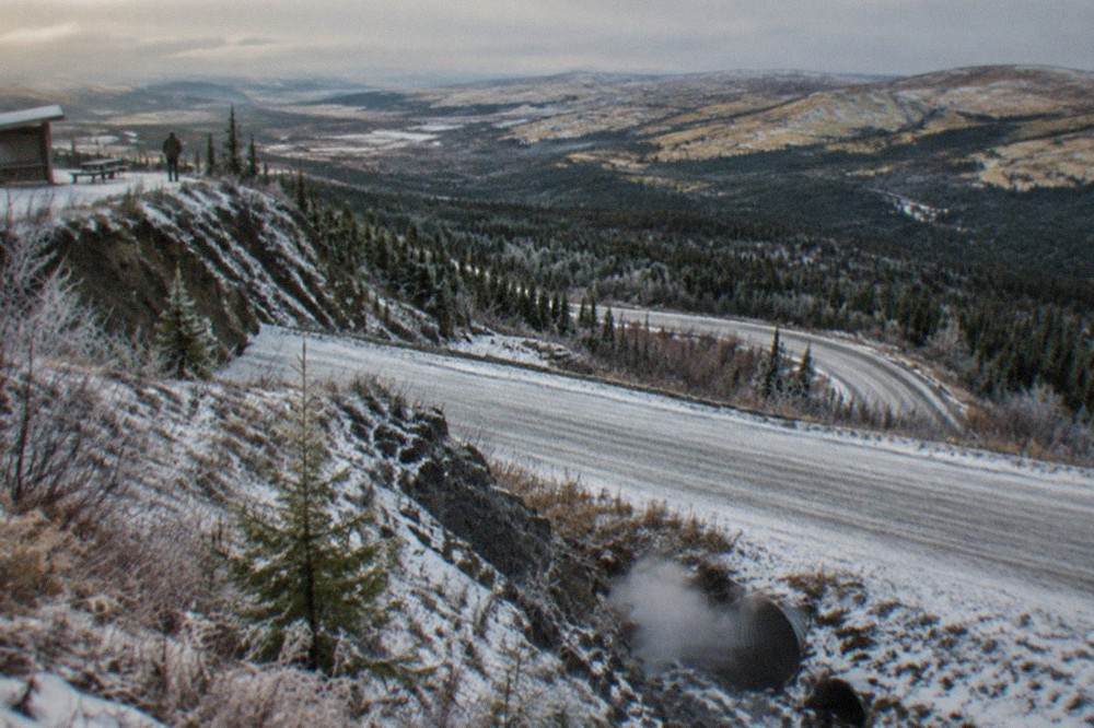 A snow-dusted gravel highway curving toward distant hills under a wide pale winter sky