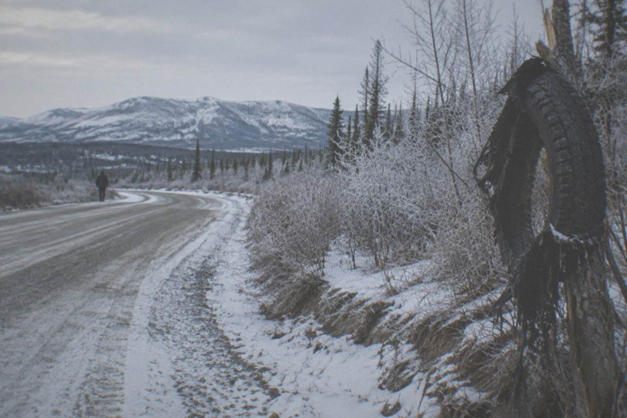 A snow-dusted gravel highway curving toward distant hills under a wide pale winter sky