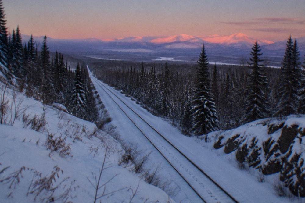 A rail line cutting through snow-covered forest with distant hills under a twilight sky