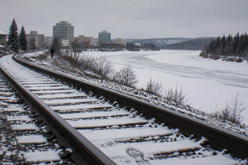 A rail line curving beside a frozen river with snow-covered hills on both sides.