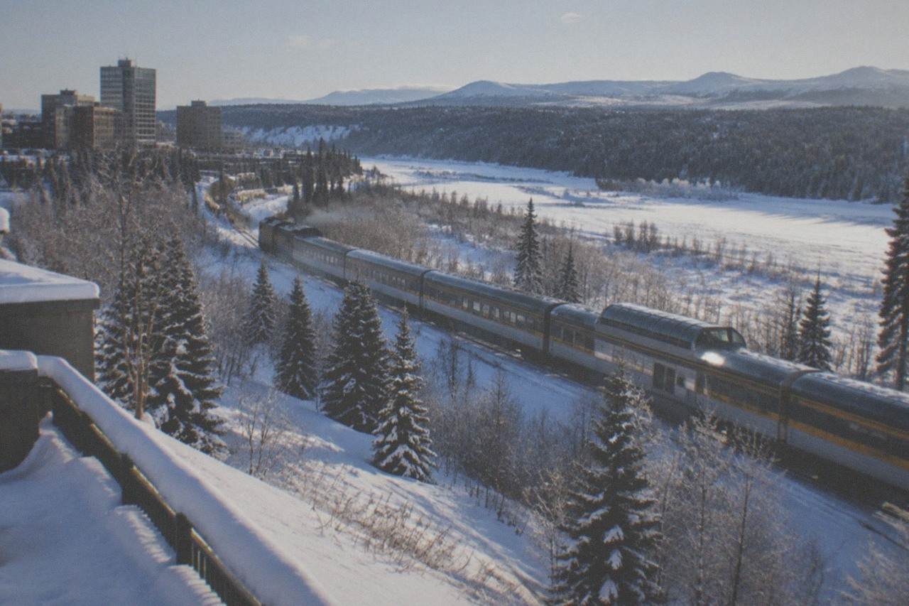 A passenger train traveling along a snow-covered Alaska river valley with distant mountains.