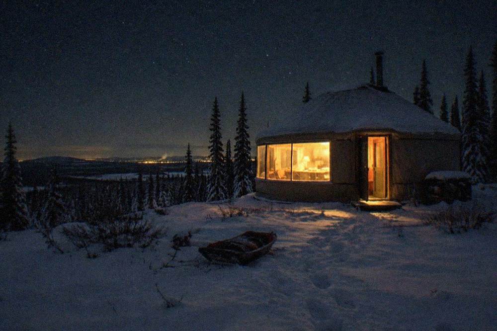 A heated yurt with a large window overlooking a dark snowy field under a star-filled sky
