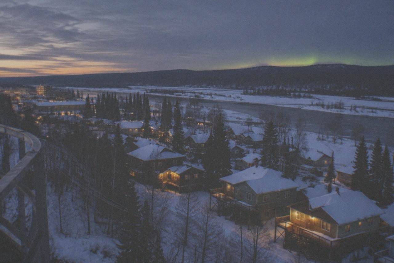 A snowy Alaska valley at dusk with distant low hills and a pale green glow beginning above the treeline.