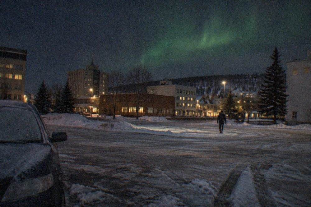 A dark winter sky above a snow-covered clearing with a faint green glow rising from the horizon