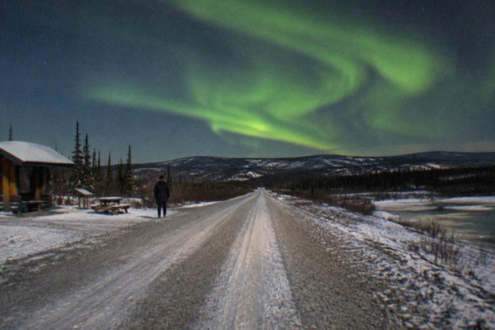 A snowy road leading toward low hills under a star-filled night sky with a green aurora band overhead