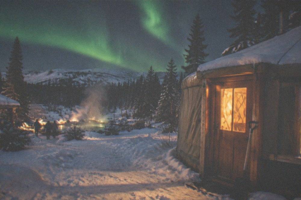 A yurt with warm windows glowing on a dark snowy landscape beneath a band of green aurora