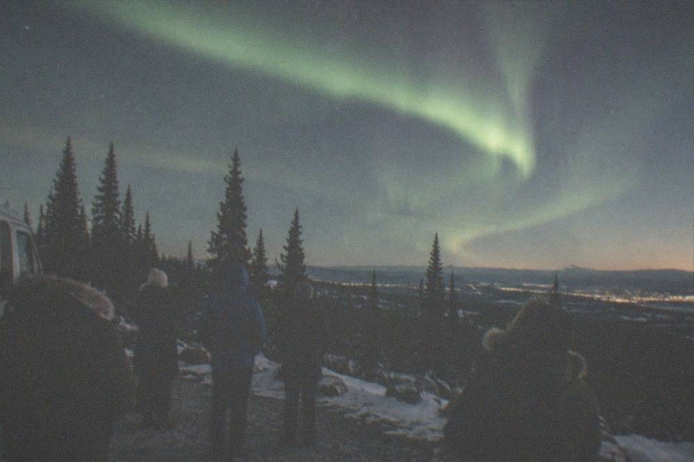 Group of visitors in winter parkers standing beside a tour van watching a green aurora arc over a dark hillside