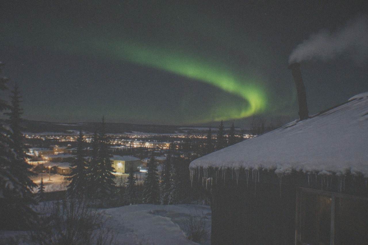 Night sky over snowy Fairbanks terrain with a bright green aurora band curving above dark spruce trees