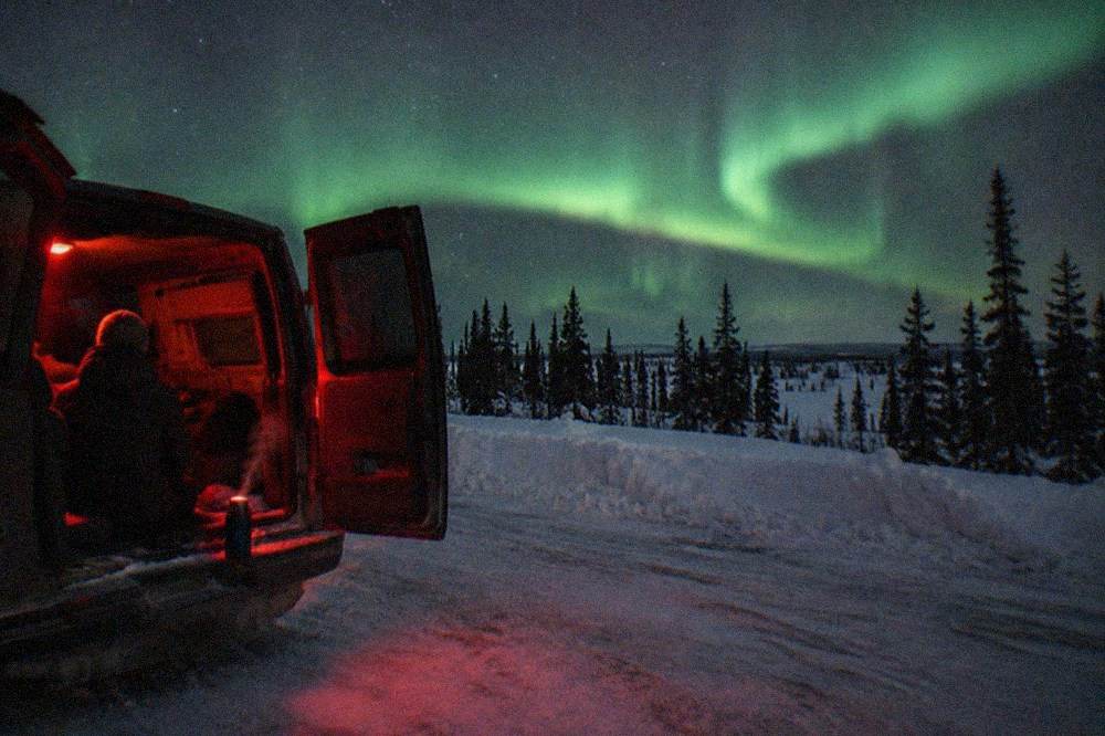 A parked tour van on a snowy roadside with green aurora bands above the treeline
