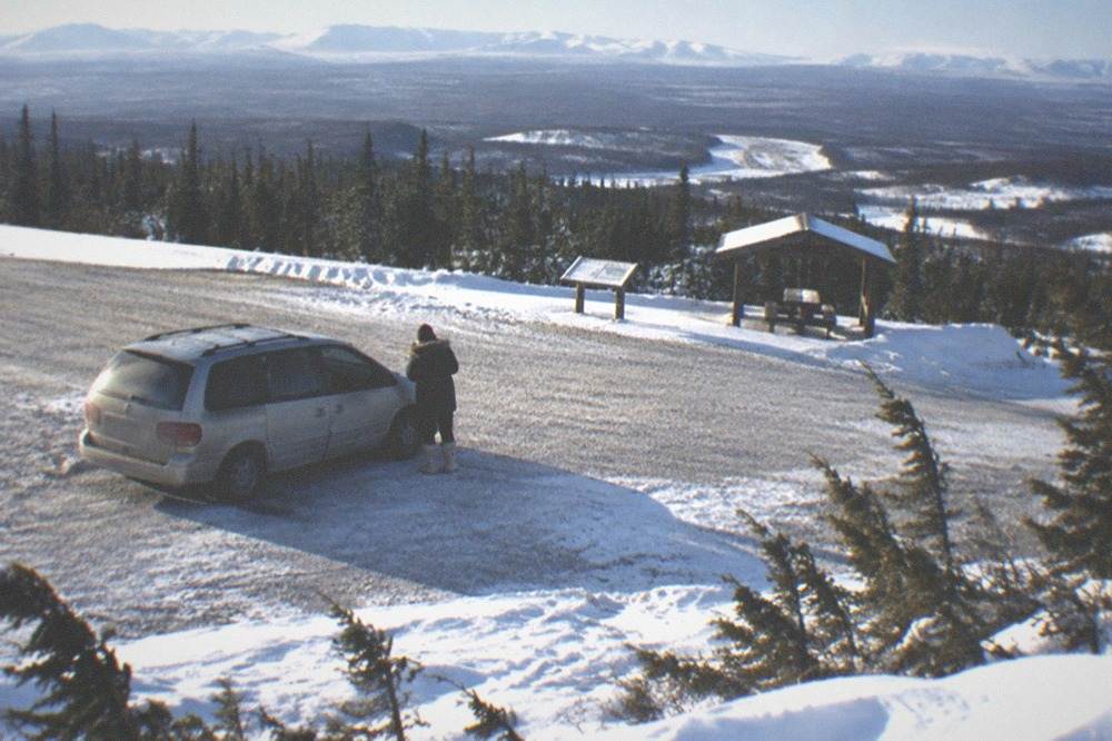 A traveler in a heavy parka and boots standing beside a van at a snowy roadside stop