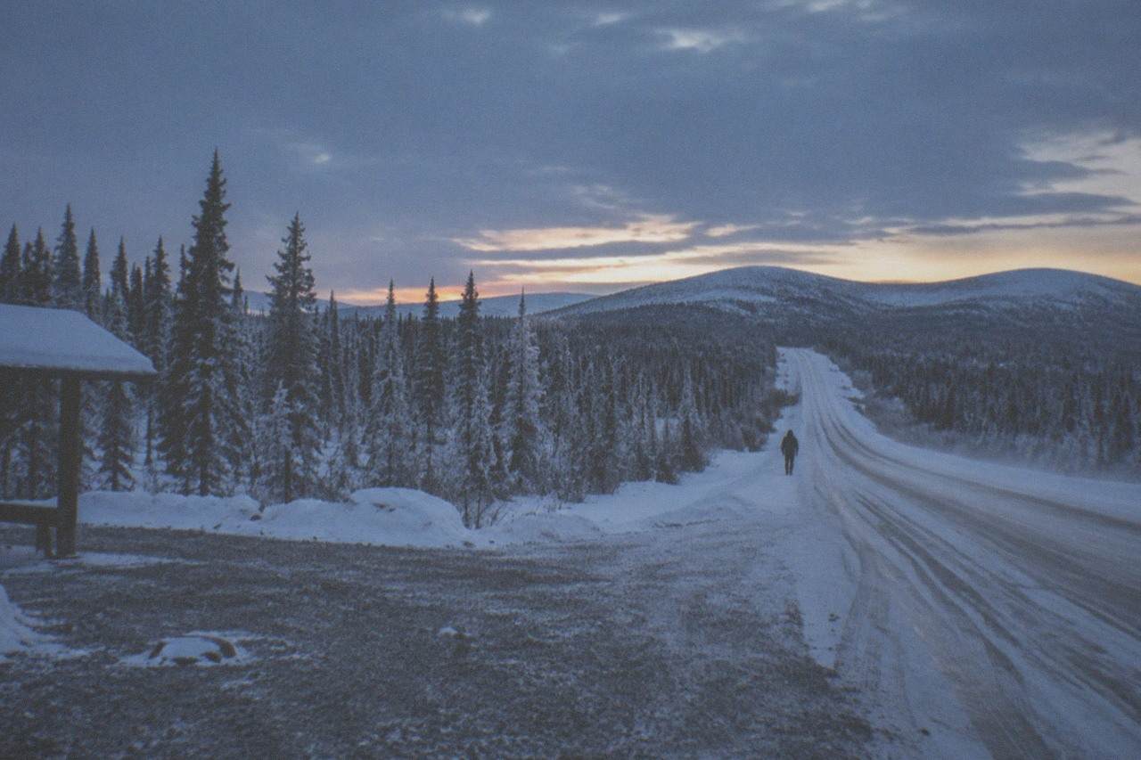 Snow-covered Dalton Highway stretching toward low hills under a dark winter sky in interior Alaska