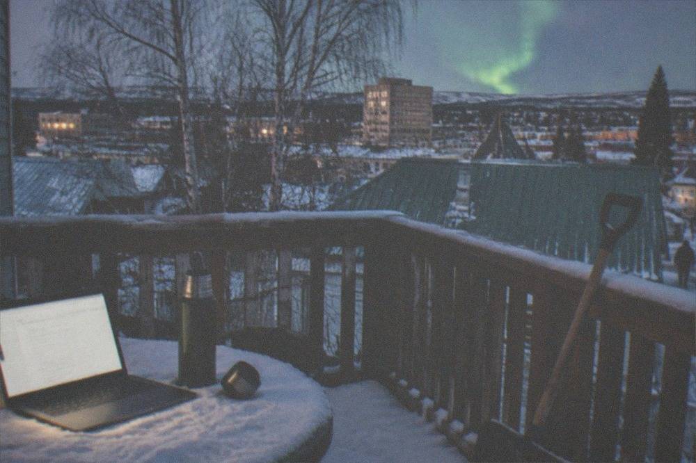 A laptop and thermos of hot drinks set up on a snowy table with a green aurora glow on the horizon behind