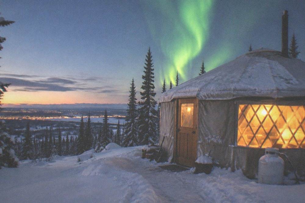 A heated yurt with a wide window and a bright aurora band glowing outside over snowy ground