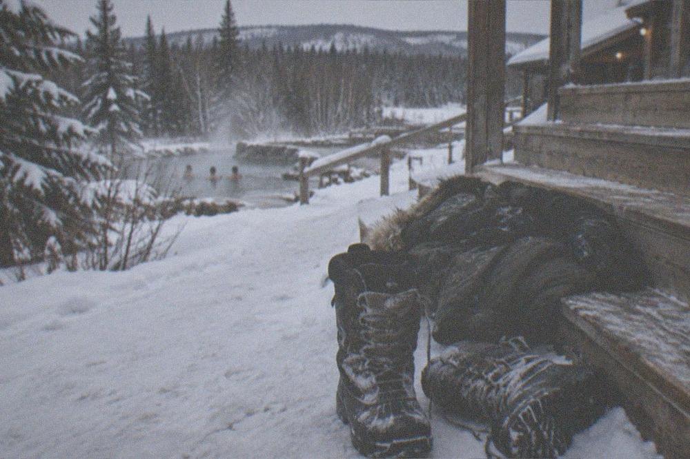 A pair of insulated winter boots standing on snow beside a parka and gloves