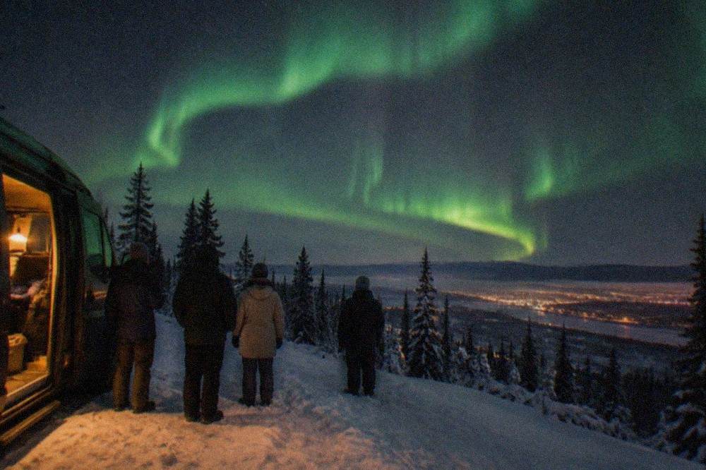 A small group of aurora watchers standing beside a heated van at a dark hilltop clearing, green aurora overhead