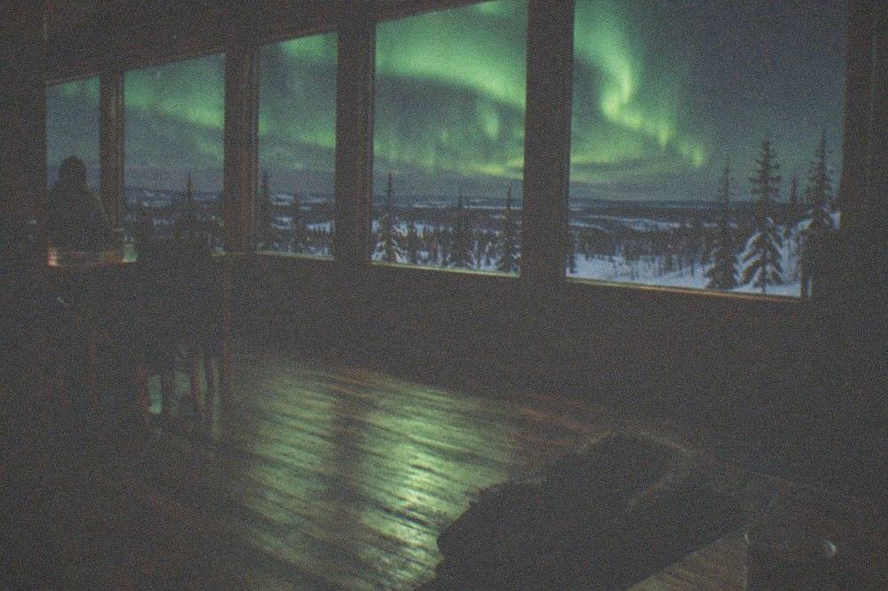 A warm timber lodge interior with large north-facing windows and green aurora light filling the glass