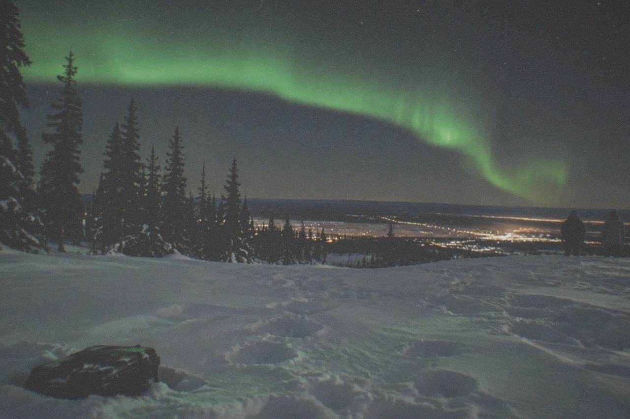 A green aurora band stretched over a snow-covered hilltop clearing outside Fairbanks under a star-filled winter sky.