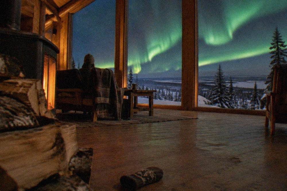 A cozy interior of a wooden aurora viewing lodge with large windows looking out over snow