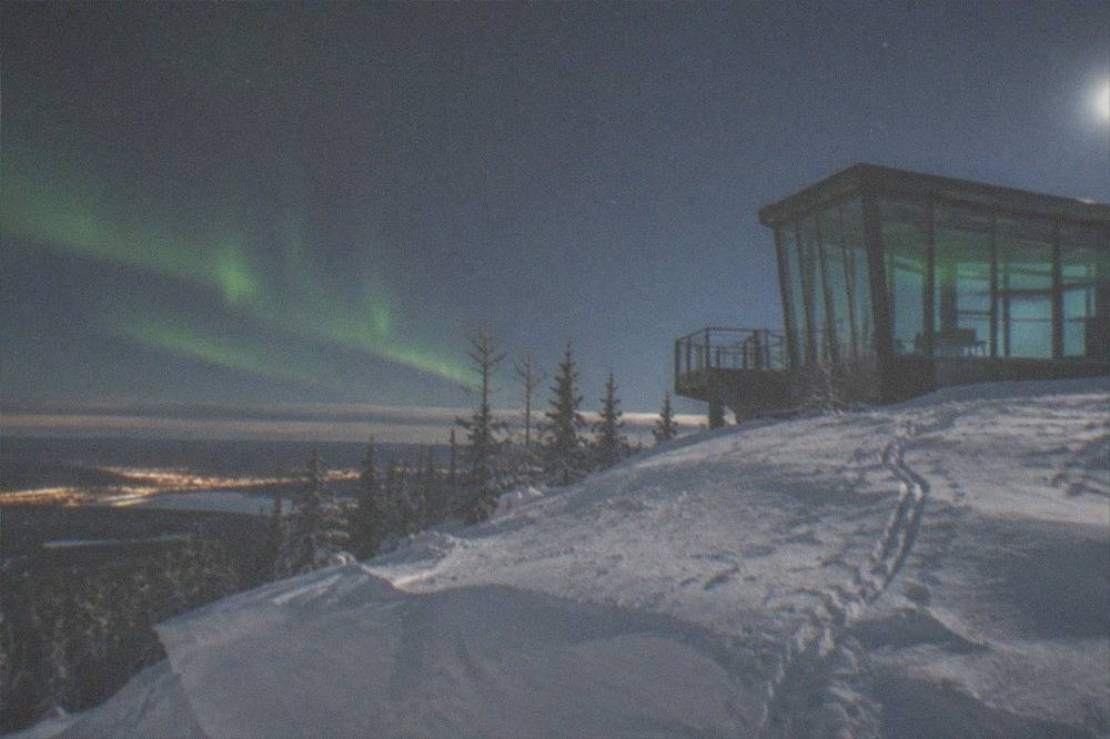 A glass-walled aurora viewing structure on a snowy summit under a starry sky