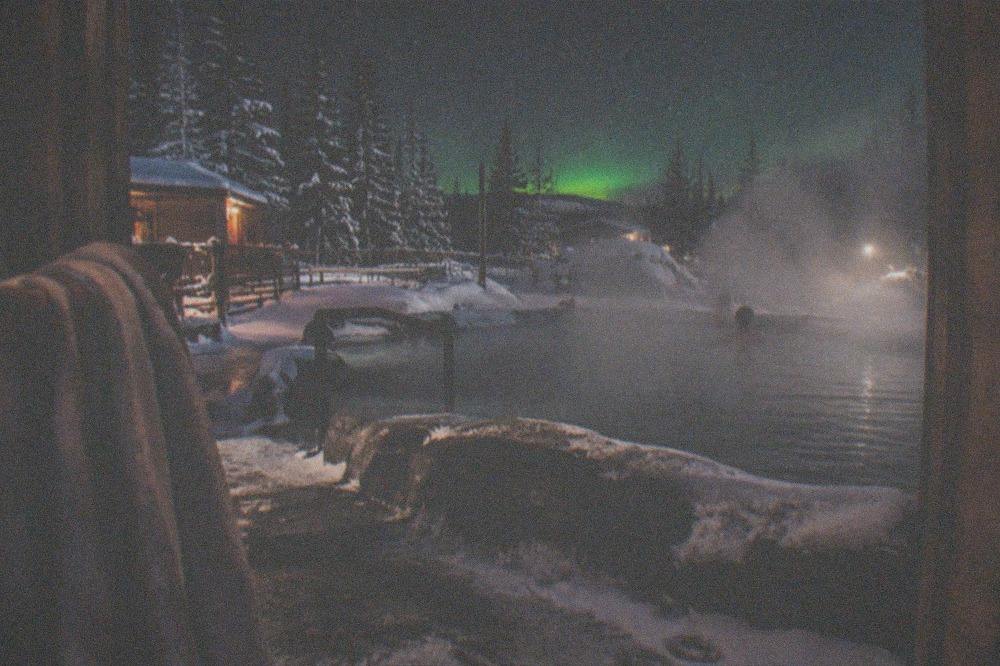 Steam rising from an outdoor hot springs pool under a dark starry sky with a faint green glow on the horizon