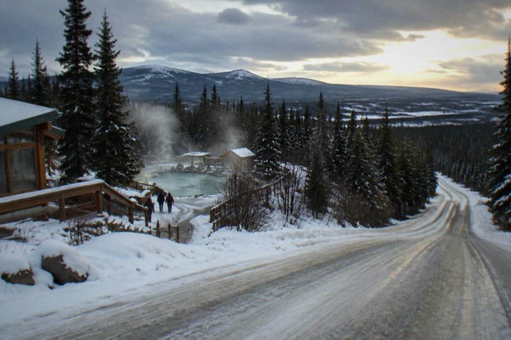 A snowy two-lane road winding through dark forest toward distant hills