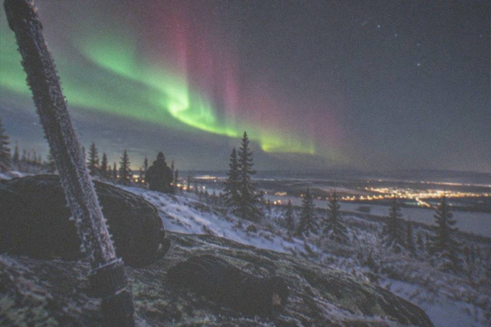 A bright green and pink aurora display over a snowy Fairbanks hillside with the Big Dipper visible