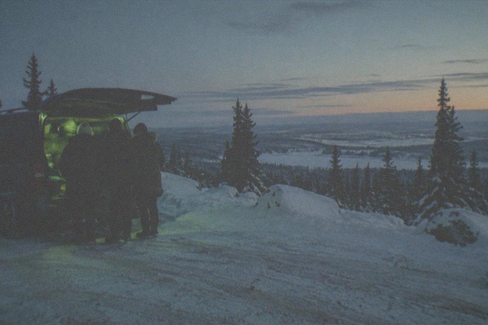 A small group of travelers in parkas standing beside a van under a dim green aurora sky near Fairbanks