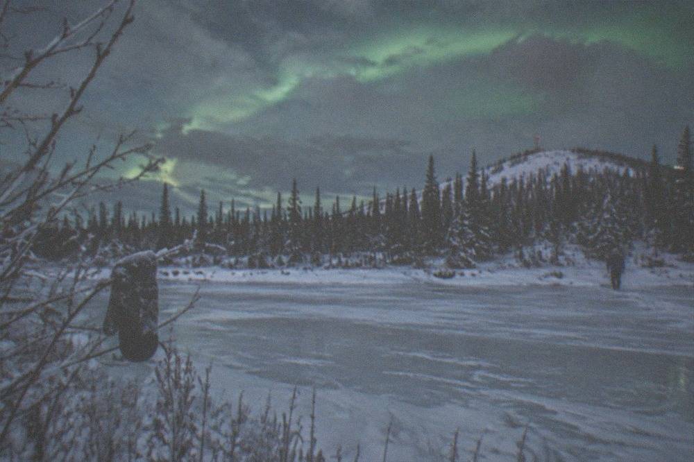 Green aurora curtain faintly visible above a snowy treeline under partly cloudy sky