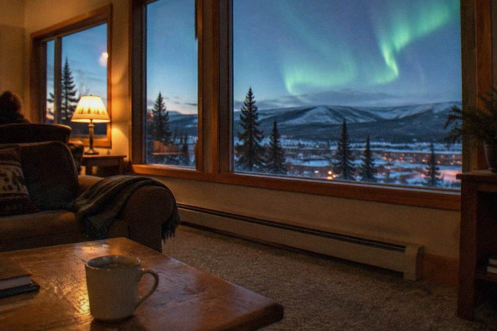 A warm lodge interior lit softly, with large windows showing a green aurora over dark hills