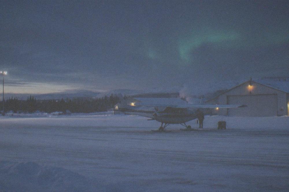 A small aircraft on a snowy runway at dusk with an aurora glow beginning in the sky above
