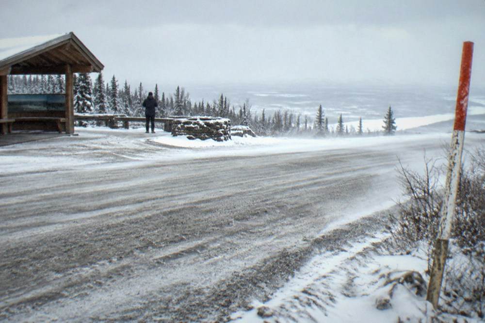 Gravel highway stretching north through flat snow-covered tundra under an overcast sky