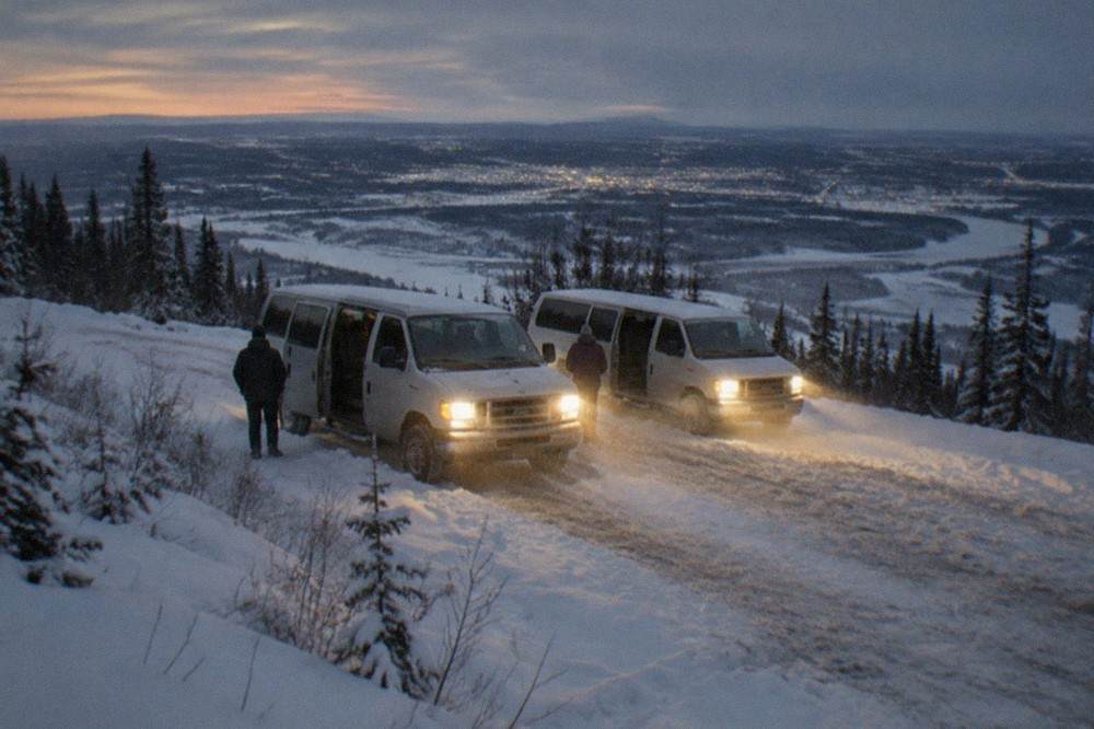 Snowy parking area at a hilltop aurora viewpoint with vans parked and headlamps glowing