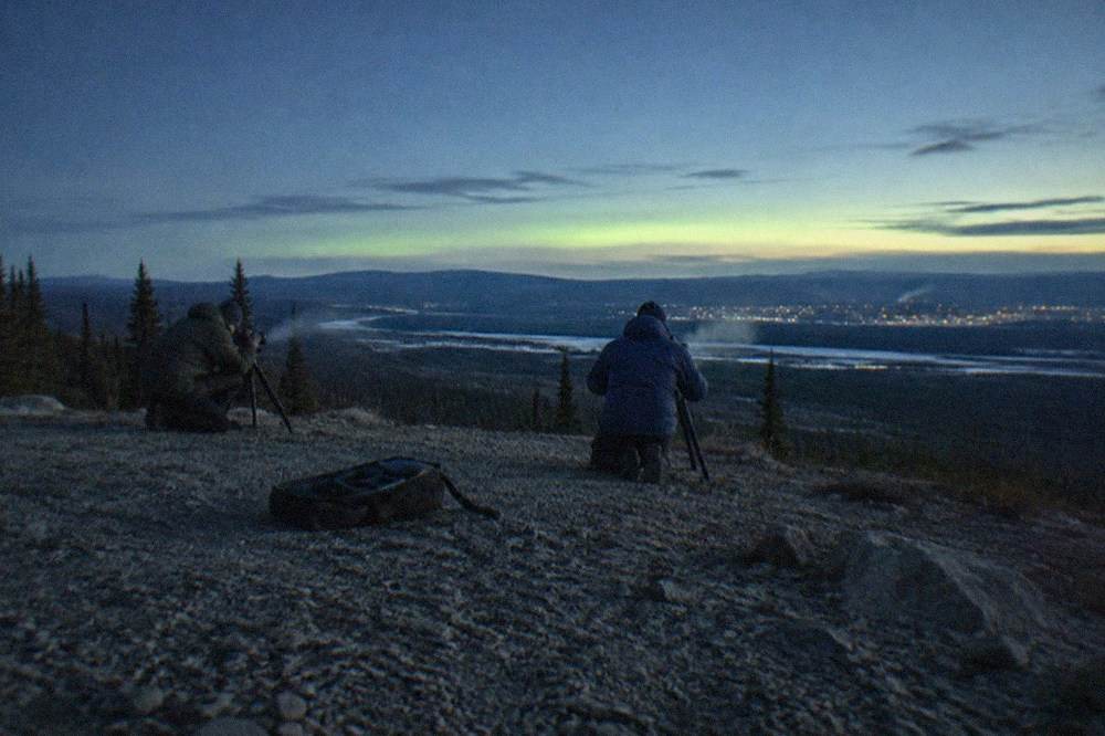Guides setting up tripods on a dark snowy hilltop while a faint green band glows on the horizon