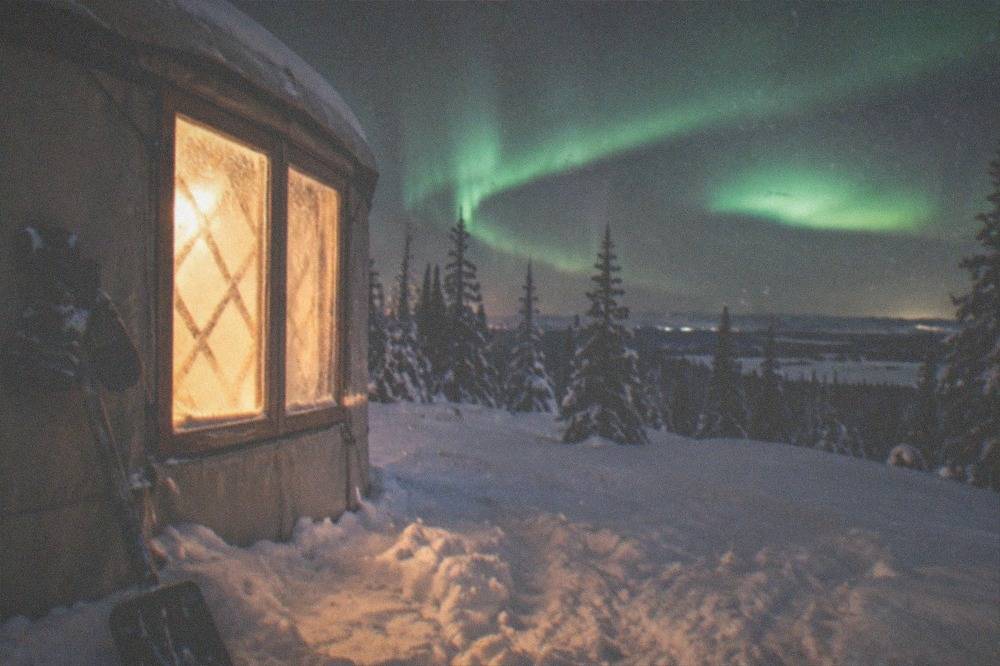 Warm glow from a heated yurt window onto the snow while a green arc stretches overhead