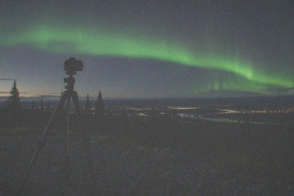 A camera on a tripod pointed at a starry sky with a green aurora band rising above the trees