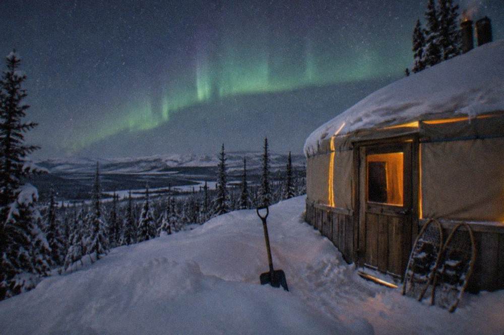 A warm wooden yurt glowing from inside under a starry sky in the hills outside Fairbanks