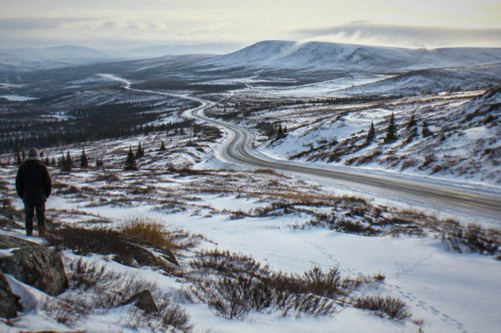 A snow-covered gravel highway winding north through open tundra hills under a pale winter sky