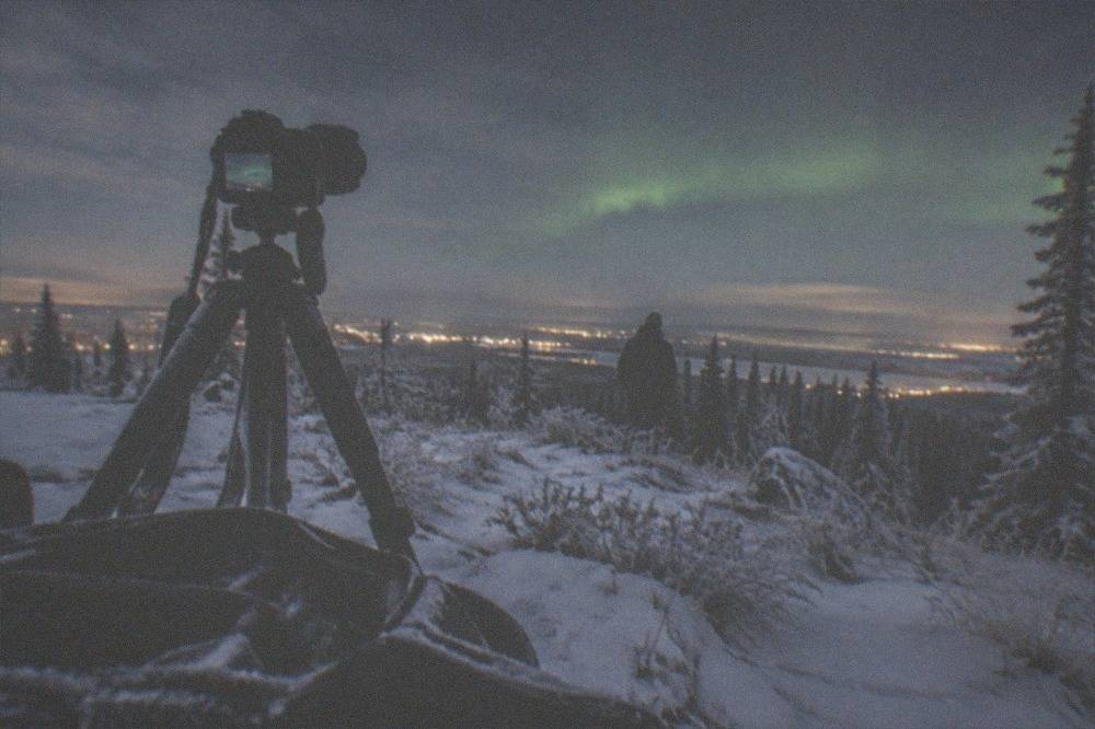 A tripod and camera bag standing on packed snow facing a faint green glow on the horizon