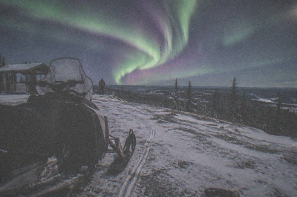 Purple and green aurora rays converging toward a point above a snow-covered hilltop