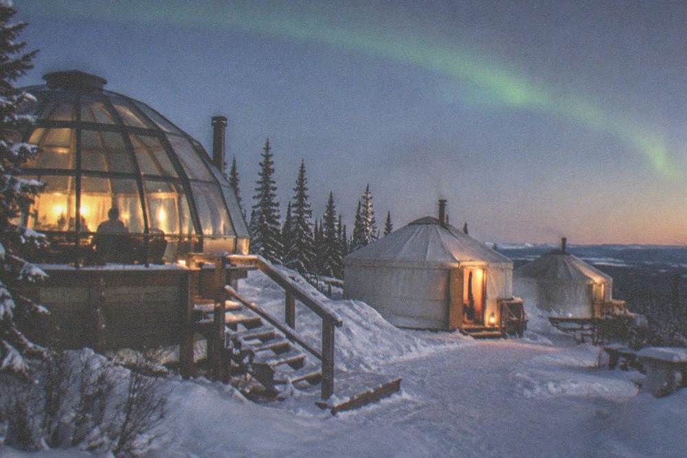 A heated dome and yurts glowing warmly on a dark hillside under a faint aurora arc