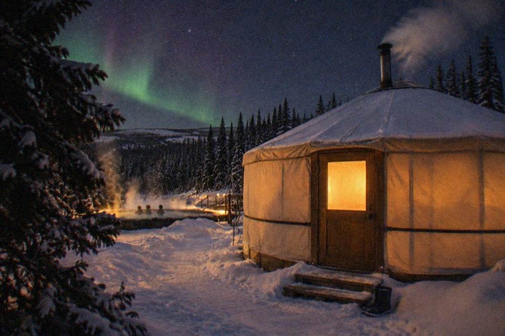 A heated yurt glowing with warm light beside a dark snowy clearing under a starry sky