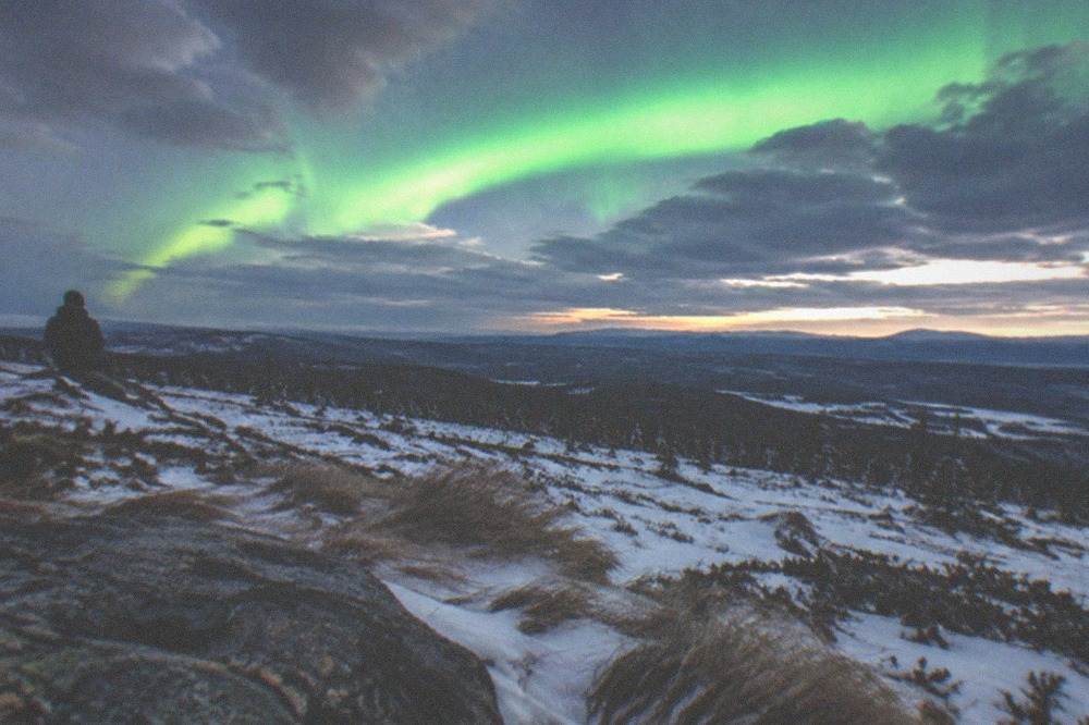 A wide snowy field under a partly cloudy night sky with a bright green aurora arc forming over distant hills