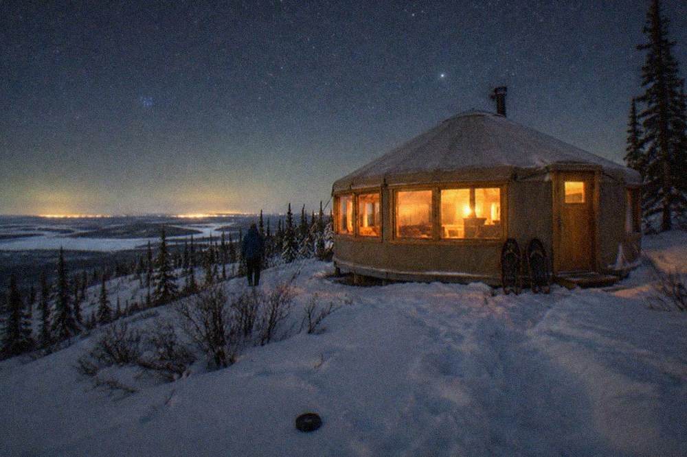 A heated yurt with large windows glowing warm under a starry sky on a snowy hillside